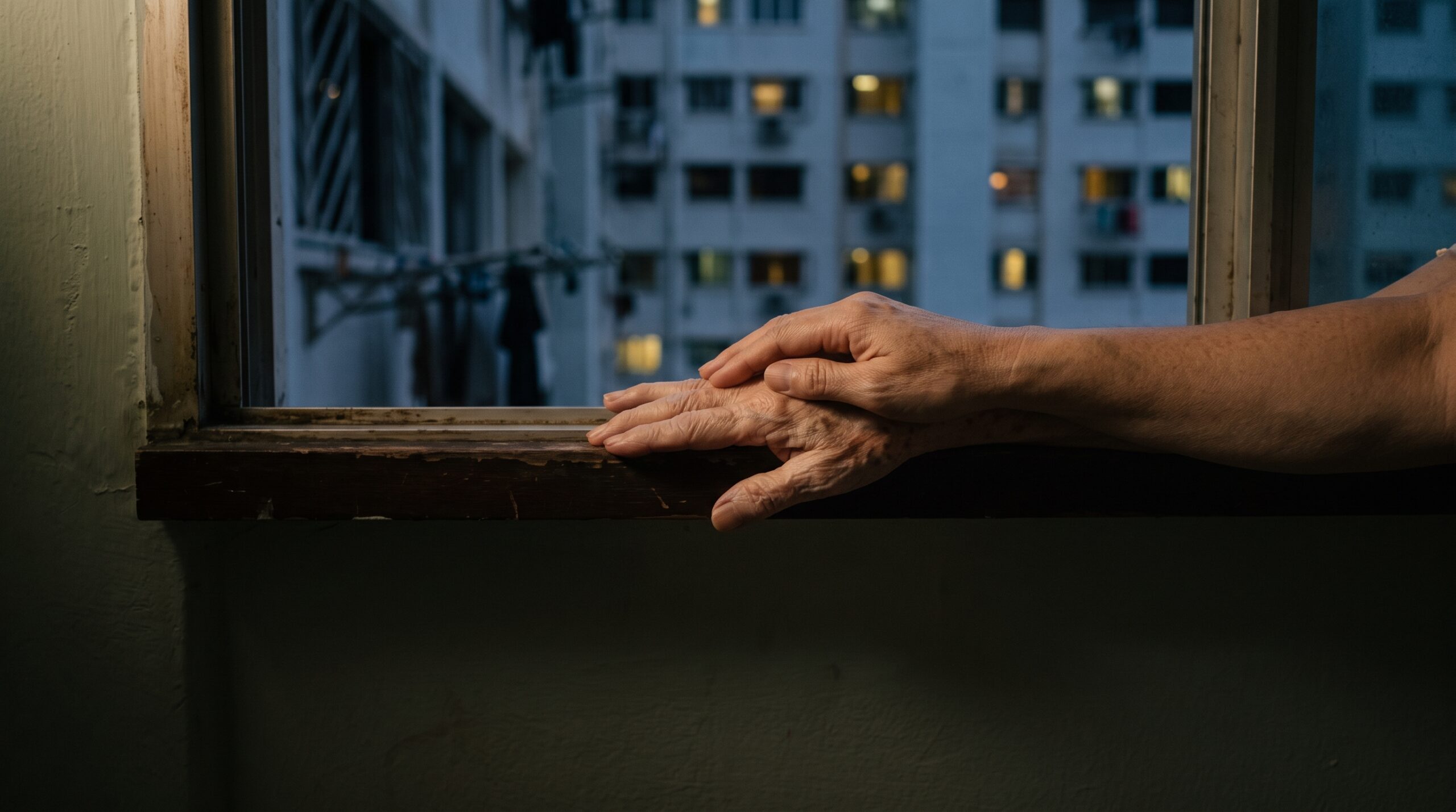 An adult child's hand resting on an elderly parent's hand on an HDB flat windowsill at dusk
